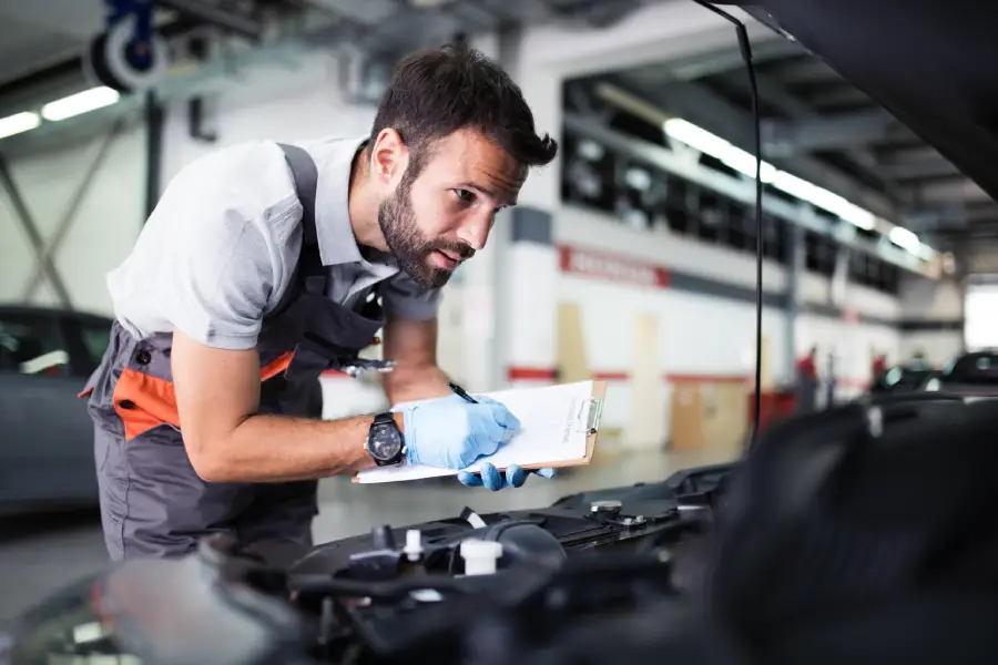 Mechanic wearing gloves and overalls inspecting a car engine while taking notes on a clipboard.