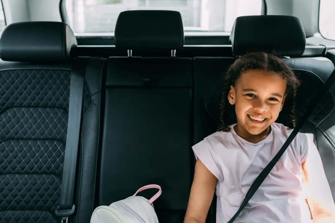 A smiling girl safely buckled into the back seat of a car, enjoying the ride.